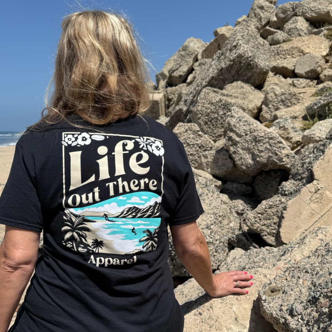 Wearing the Unisex Aloha Tee in black by Life Out There Apparel, a person with long hair stands on rocky beach terrain on Oahu’s North Shore; the shirt features a tropical beach graphic on the back.