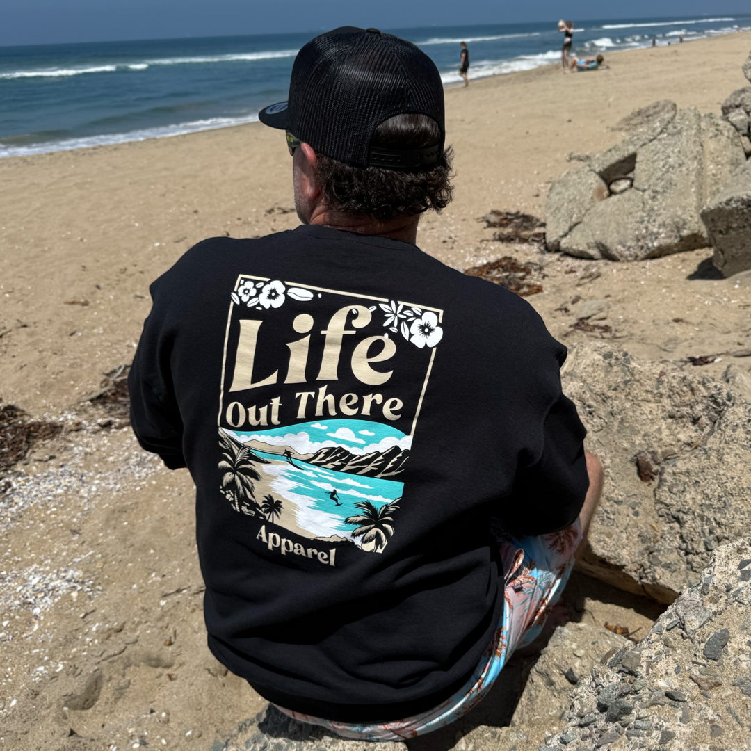 A man in a black Unisex Aloha Crew Neck Sweatshirt by Life Out There Apparel sits on rocks at a sandy North Shore Oahu beach, facing the ocean with a few people in the distance, capturing true Hawaiian vibes.