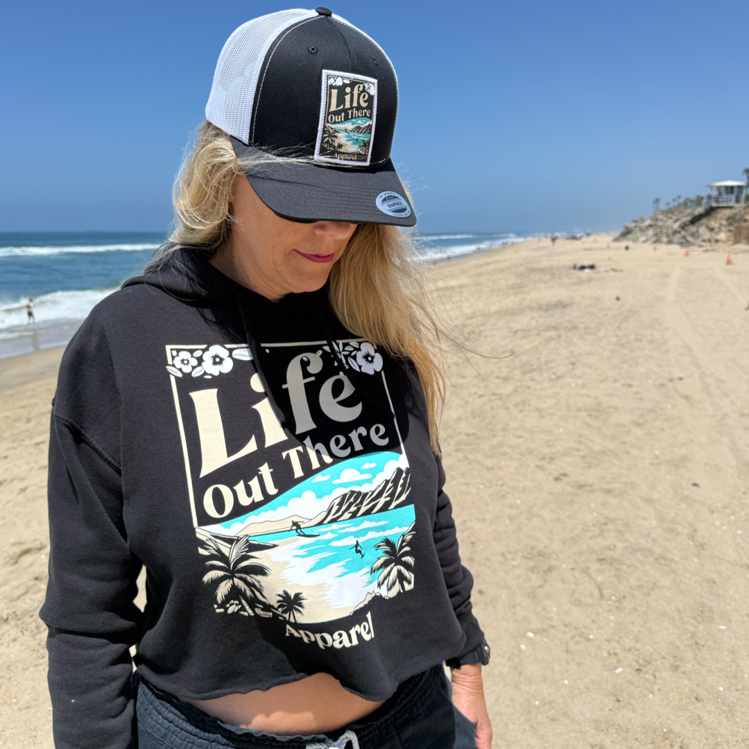 A woman wearing a Life Out There Apparel hat and the Womens Aloha Crop Hoodie in black stands on a sunny North Shore beach in Oahu, with ocean waves and distant houses behind her, embodying true Hawaiian vibes.