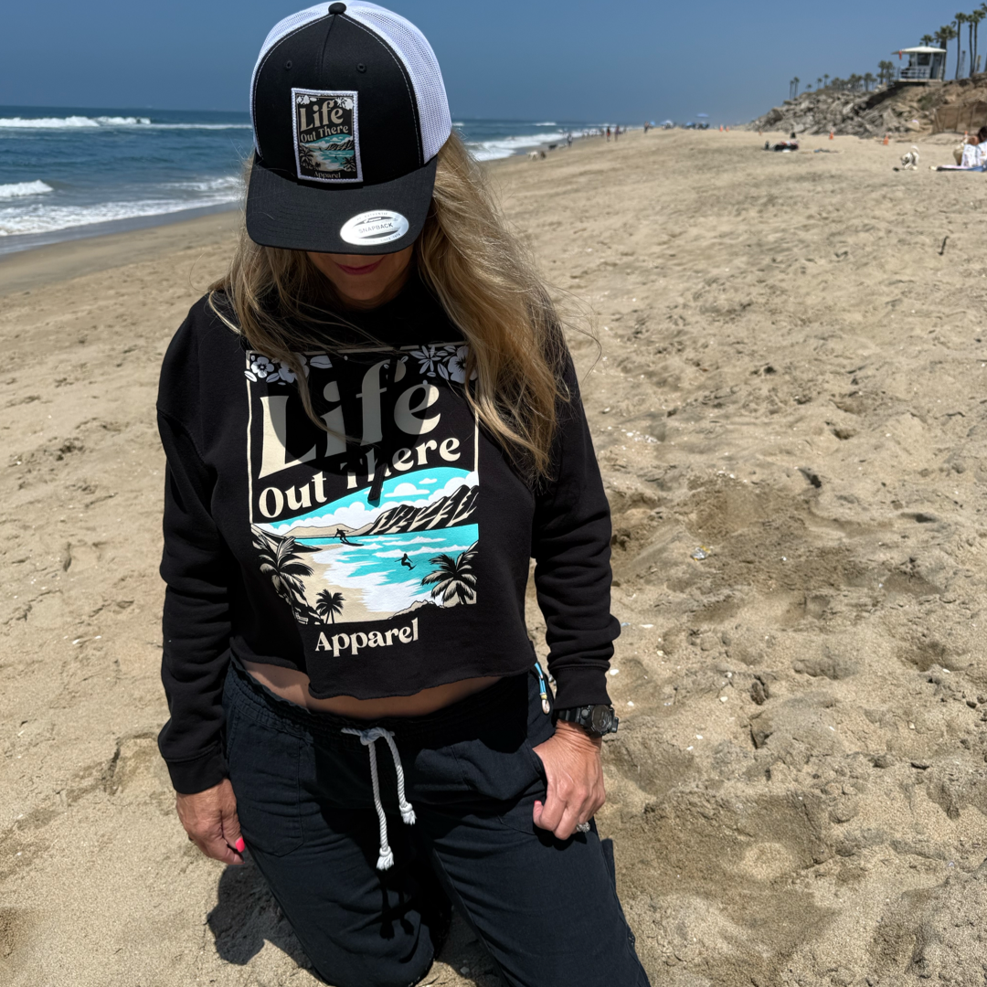 A woman stands on a sandy North Shore beach wearing the Womens Aloha Crop Hoodie in black from Life Out There Apparel, plus a matching black-and-white hat. She looks down, with ocean waves and blue sky creating Hawaiian vibes.