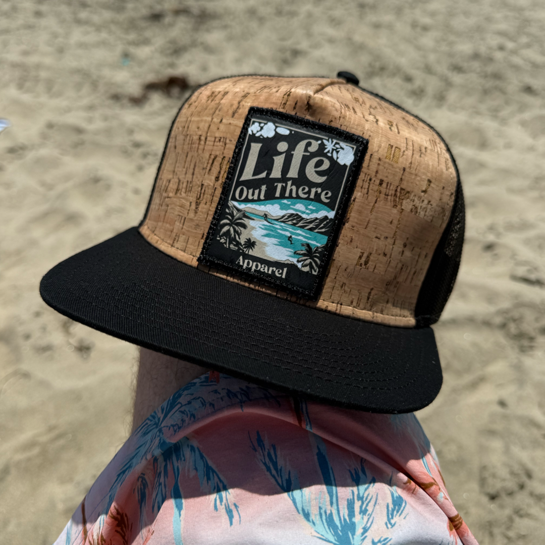A tan cork and black ALOHA Flat Bill SnapBack Trucker Hat by Life Out There Apparel sits on a knee at the beach, paired with pink and blue palm leaf shorts, against North Shore Oahu sand, giving off Hawaiian vibes.