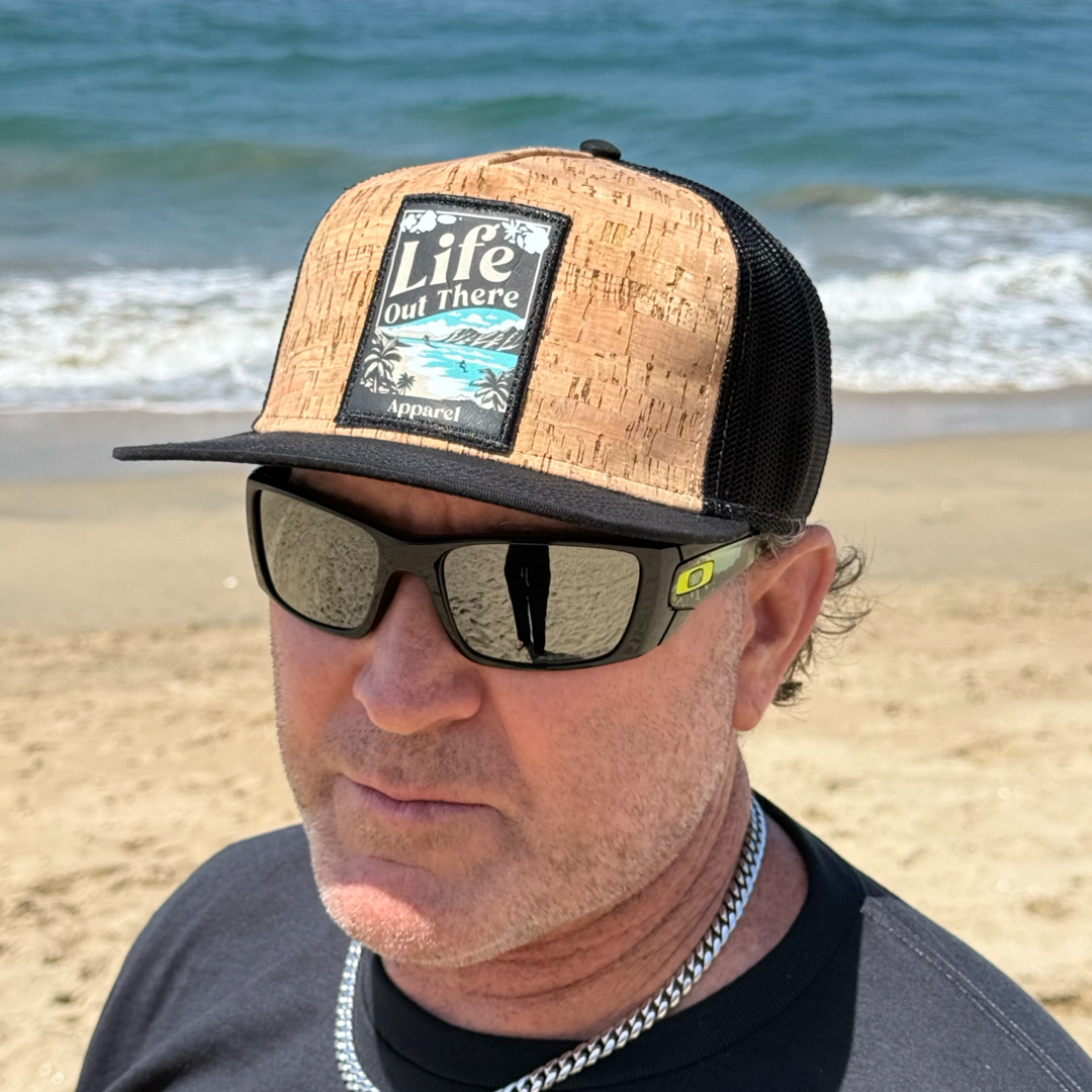 A man wearing the ALOHA - Flat Bill SnapBack Trucker Hat (Cork/Black) by Life Out There Apparel and dark sunglasses stands on a sandy beach with the ocean behind him, radiating Hawaiian vibes.