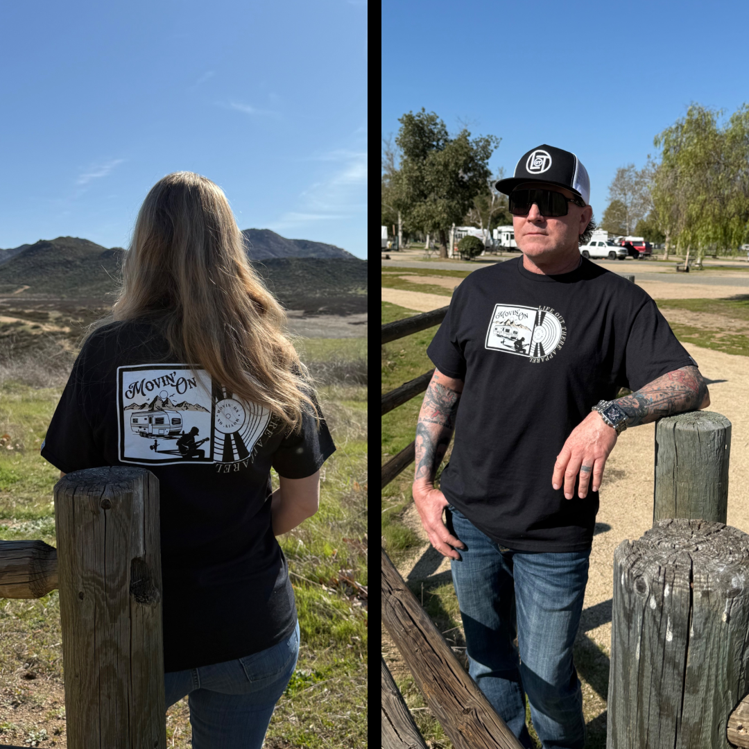 Two people outdoors wear the Unisex - Movin On Tee - Black by Life Out There Apparel; one shows the back design with a camper, vinyl record, and album cover theme, while the other stands facing forward by a wooden fence.