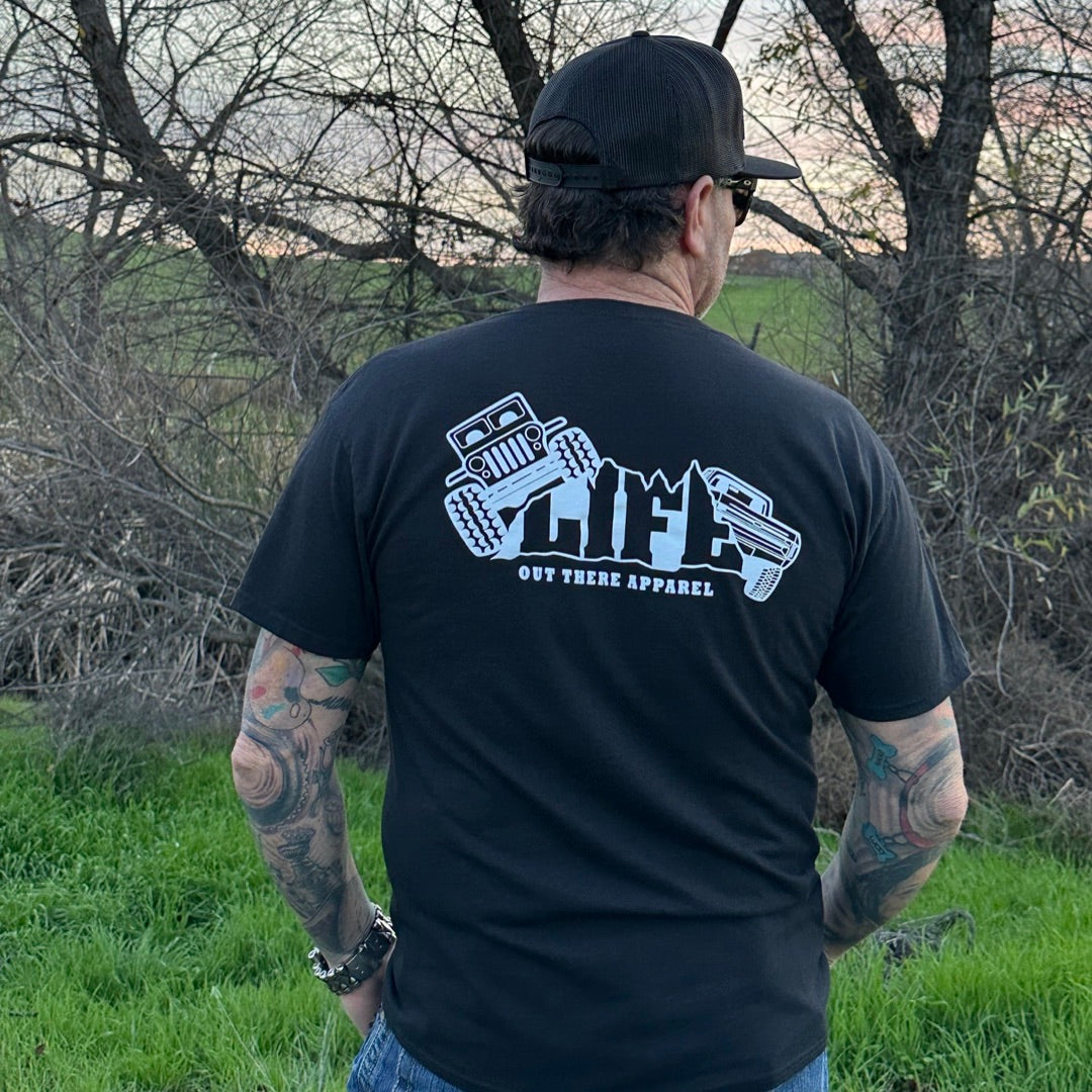 A tattooed man, dressed in the Unisex The Crawler T Shirt - Black by Life Out There Apparel and a black cap, stands on grass with trees in the background. Facing away, he imagines an off-roading adventure while enjoying the scenic view of nature.