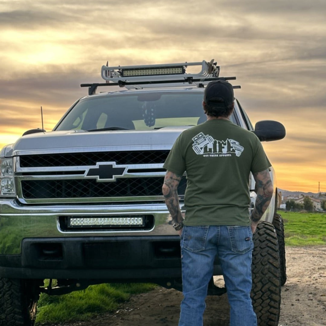 Wearing the Unisex The Crawler T-Shirt in Military Green from Life Out There Apparel, a person stands in front of a robust pickup truck designed for off-roading, complete with off-road tires and a roof rack. Against the backdrop of a magnificent sunset sky, this adventurous scene comes to life as they sport jeans and a hat.
