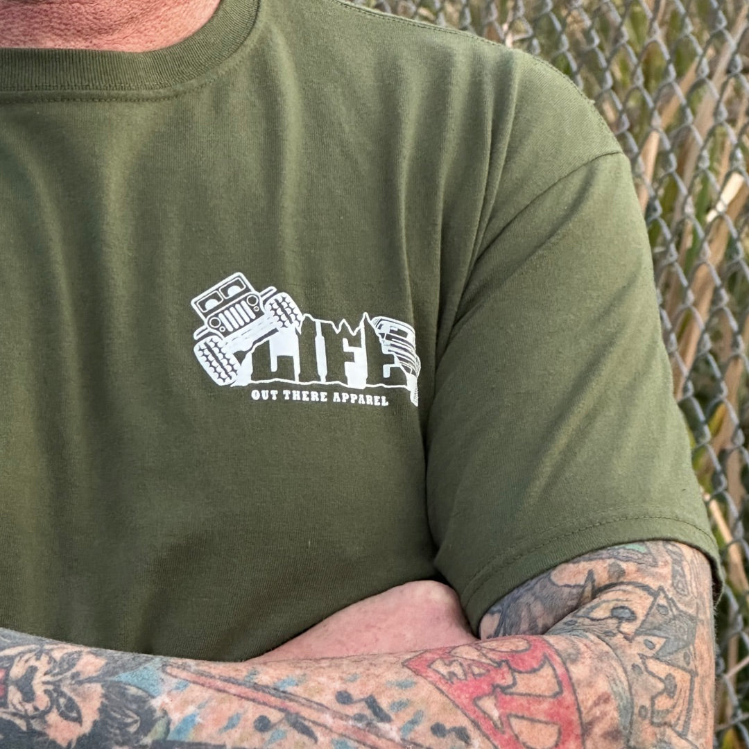A person with tattooed arms confidently crosses their arms while wearing a Unisex The Crawler T Shirt in Military Green from Life Out There Apparel. Featuring a logo with a jeep and a dirt bike, they stand poised for their next off-roading adventure in front of a chain-link fence.