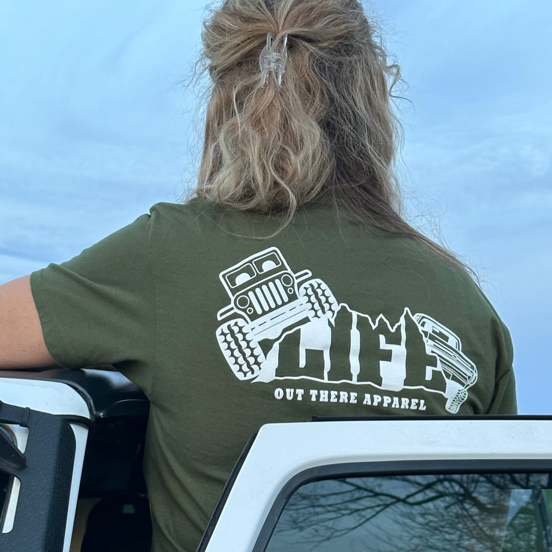 A person with long hair stands next to an open Jeep door, wearing a Life Out There Apparel Unisex The Crawler T-Shirt in Military Green, which features a Jeep graphic. The clear blue sky hints at thrilling off-roading adventures beckoning beyond the horizon.