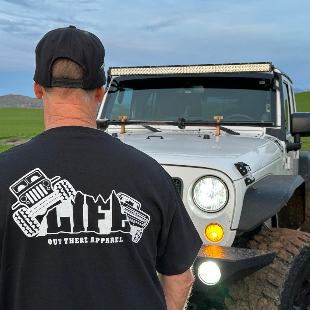 A person dressed in a Life Out There Apparel Unisex The Crawler T Shirt - Black poses in front of a white off-road vehicle designed for rock crawling, featuring rugged tires and a roof light bar, set against a backdrop of green fields and a blue sky speckled with clouds.