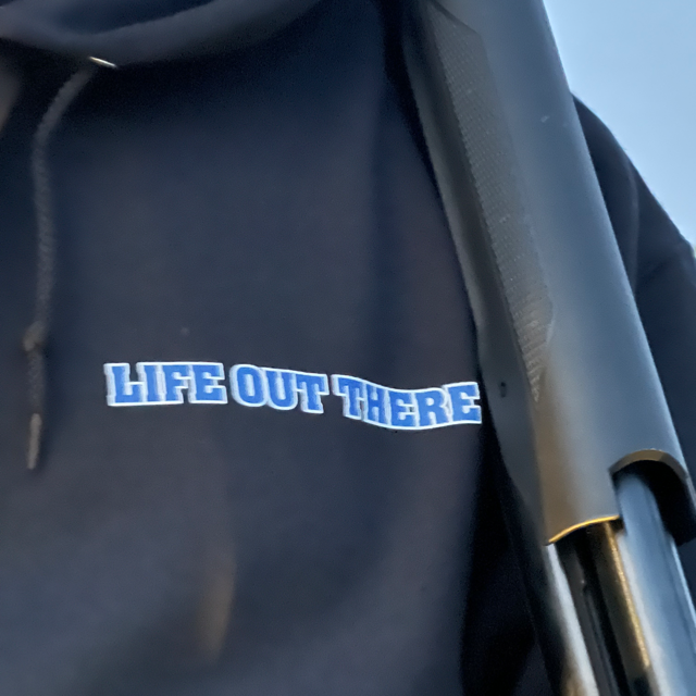 A close-up image features a person wearing the Unisex The Freedom Hoodie - Black from Life Out There Apparel, which prominently displays the words LIFE OUT THERE in blue and white lettering. They are holding an object with a black handle.