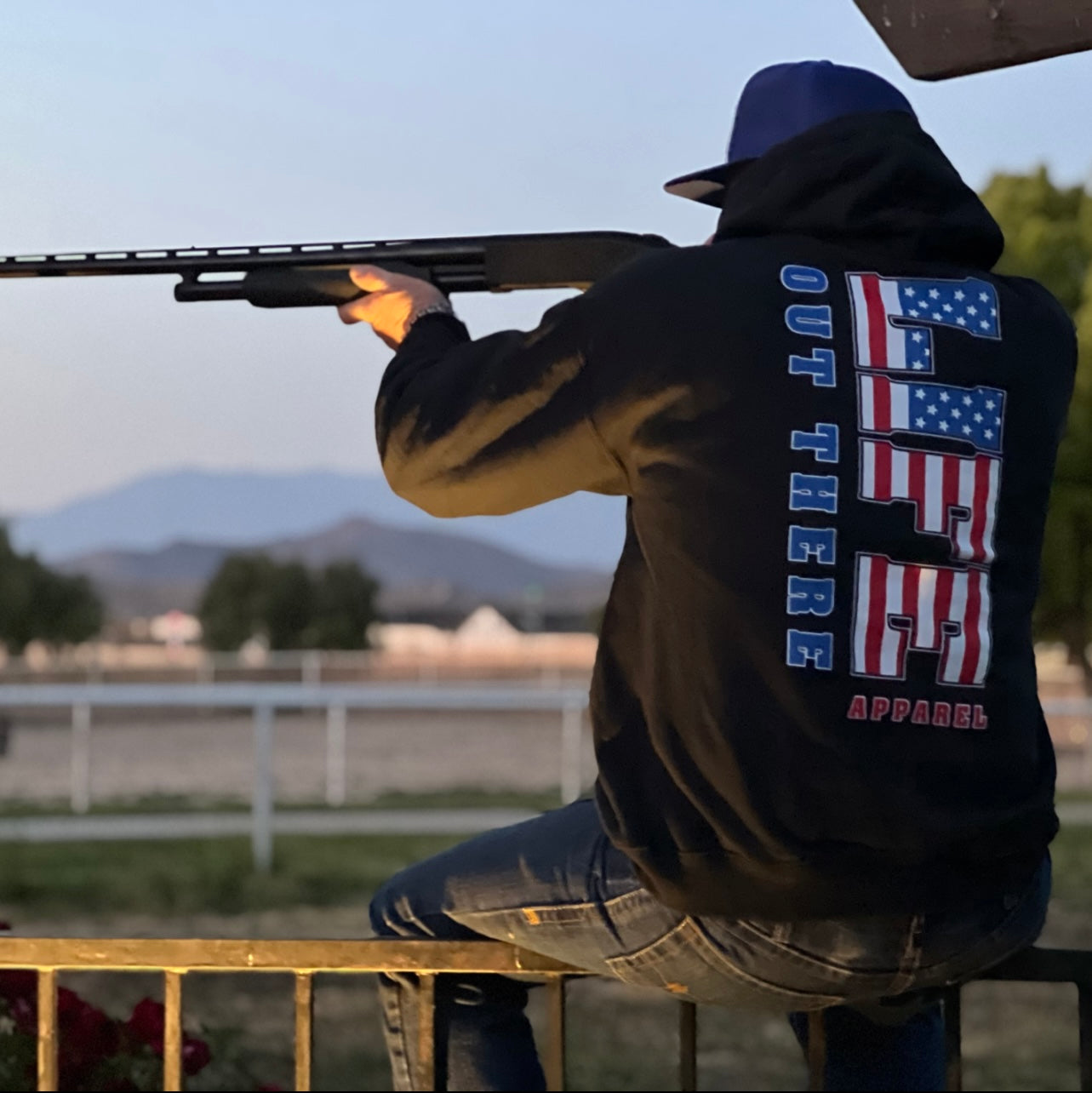 Clad in the Unisex The Freedom Hoodie - Black from Life Out There Apparel, an individual aims a rifle outdoors, epitomizing the essence of The Freedom. Surrounding them are a fence, trees, and mountains beneath the clear dusk sky, evoking the spirit of the Land of the Free.