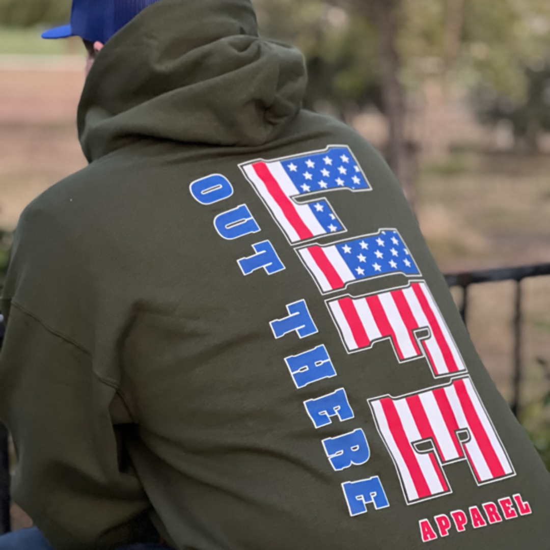 A man wears the Unisex The Freedom Hoodie in military green, adorned with CFT styled like the American flag to celebrate the spirit of freedom. Below, it proudly displays Life Out There Apparel. The background is a gently blurred outdoor scene.