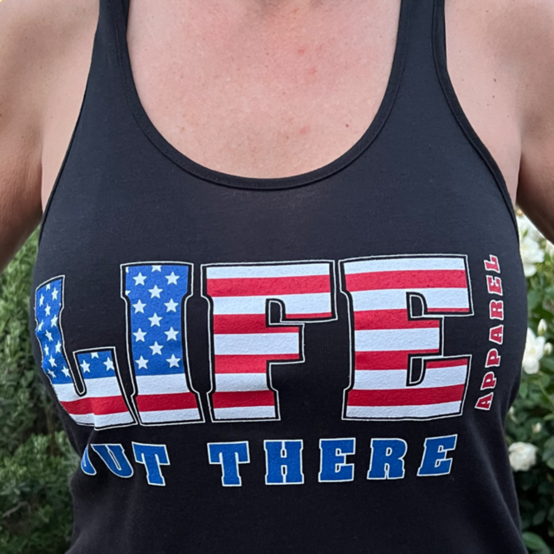 A person is wearing "The Freedom Women's Tank Top - Black" from Life Out There Apparel, featuring the word LIFE in large letters filled with an American flag pattern, ideal for patriotic apparel fans. Underneath, it reads OUT THERE in blue letters with the word APPAREL written vertically on the side, making it perfect for a 4th of July release.