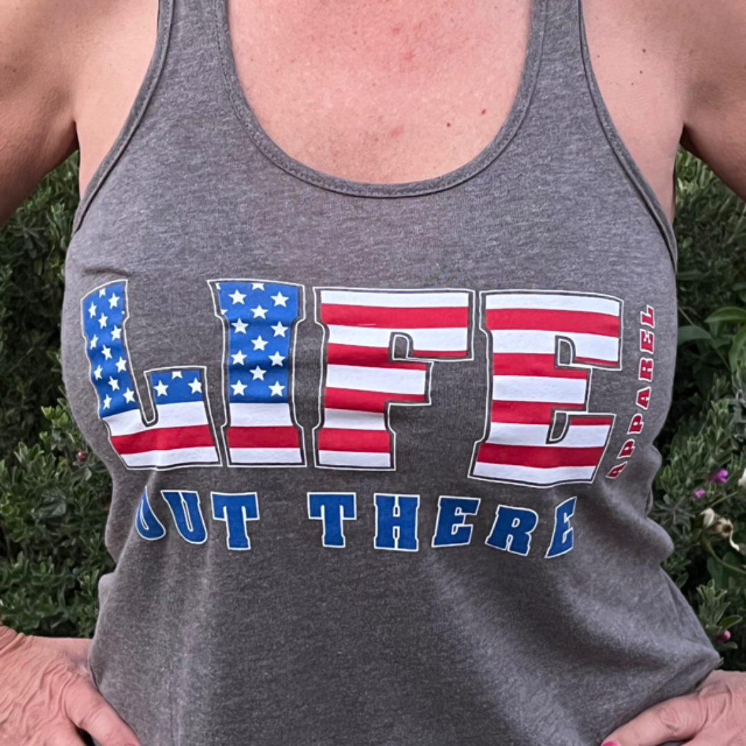 A close-up features a person wearing The Freedom Women's Tank Top - Grey by Life Out There Apparel, adorned with the word "LIFE" in a pattern resembling the American flag for a patriotic touch. Below it reads "OUT THERE" in blue text. The person stands with hands on their hips against a vibrant, green background.