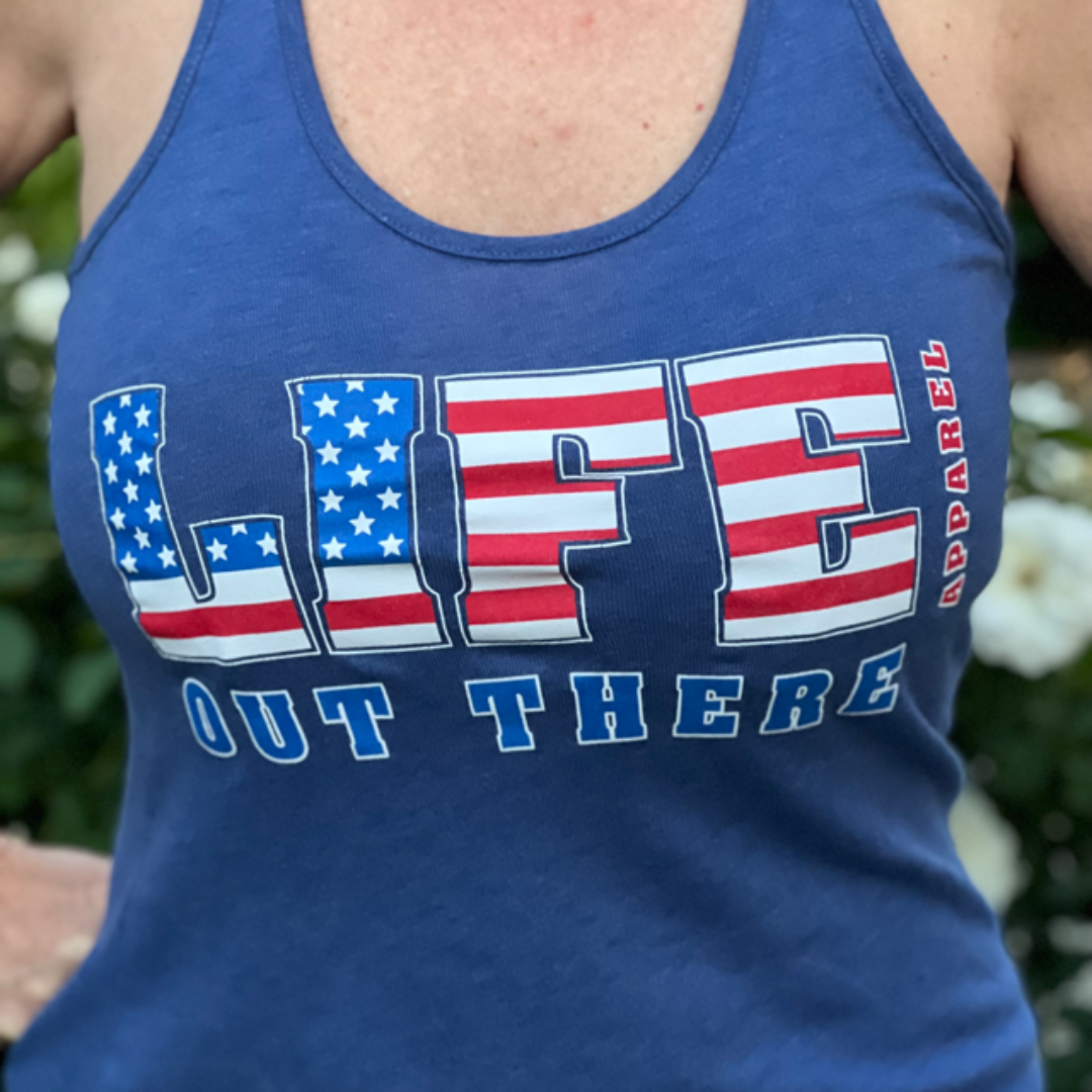 A person wearing The Freedom Women's Tank Top in Royal from Life Out There Apparel stands out with "LIFE" designed in an American flag pattern, adding a patriotic touch. White flowers and greenery flourish in the background, enhancing the scene's vibrant embrace of freedom.