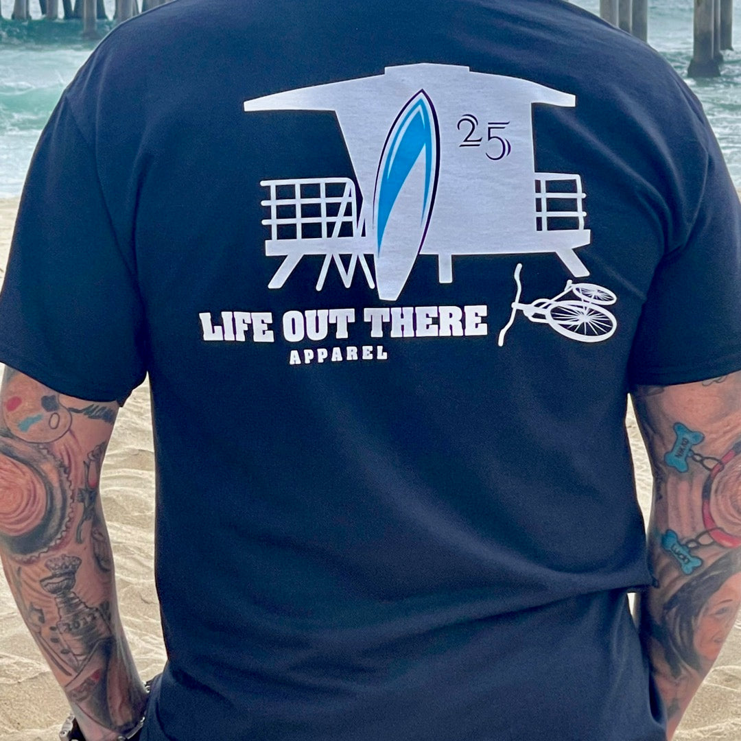 A person with tattooed arms stands on a beach wearing a black Unisex The JJ T Shirt, featuring a lifeguard tower, surfboard, and bicycle graphic. The design highlights the brand Life Out There Apparel printed below. Ocean waves and pier pilings create the perfect backdrop for this summer vacation scene.