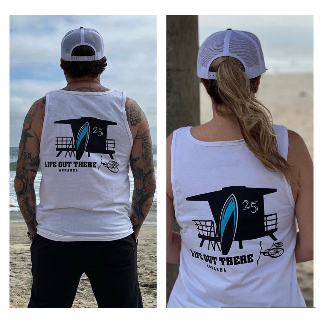 Two individuals wearing the Unisex The JJ Tank Top - White from Life Out There Apparel, which features a black and blue surfboard and lifeguard tower design, are standing on the beach. One wears a black cap while the other sports a white one, with ocean waves and sandy shoreline in the background.