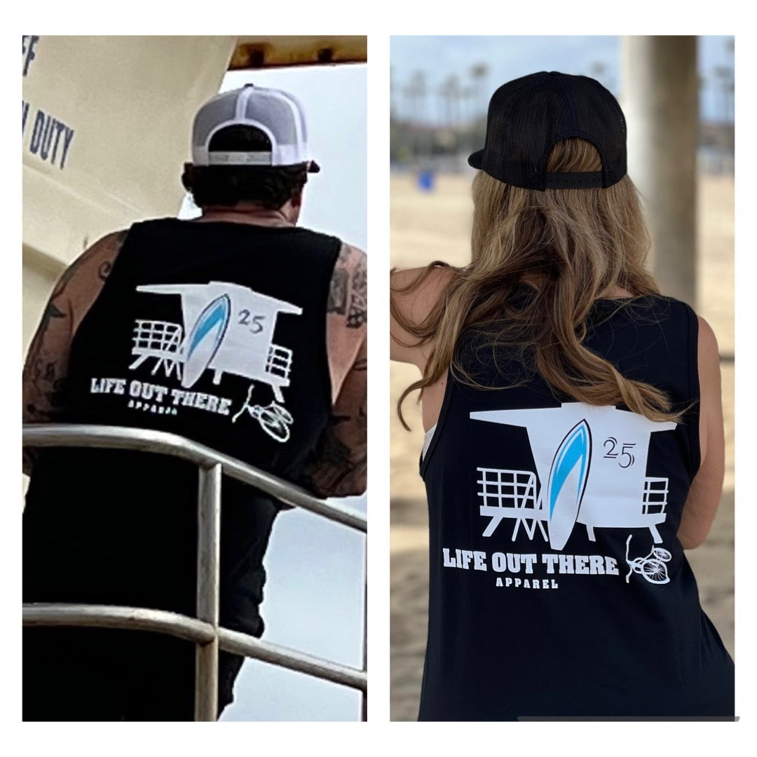 Two individuals wearing Unisex The JJ Tank Top in black, along with baseball caps, stand on the beach. Their shirts feature a lifeguard tower graphic numbered 25 and the brand name "Life Out There Apparel." Beach cruisers and pier elements in the background enhance the quintessential coastal vibe of the scene.