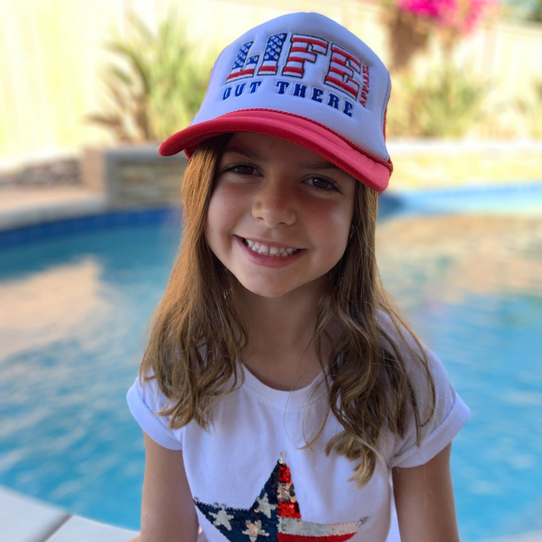 A smiling young girl with long brown hair stands by a pool, sporting a red and white "Youth The Freedom Embroidered Snapback Trucker Hat" from Life Out There Apparel, along with a white T-shirt showcasing a sequined American flag star design—ideal American attire for celebrating the 4th of July. The background features greenery and water.