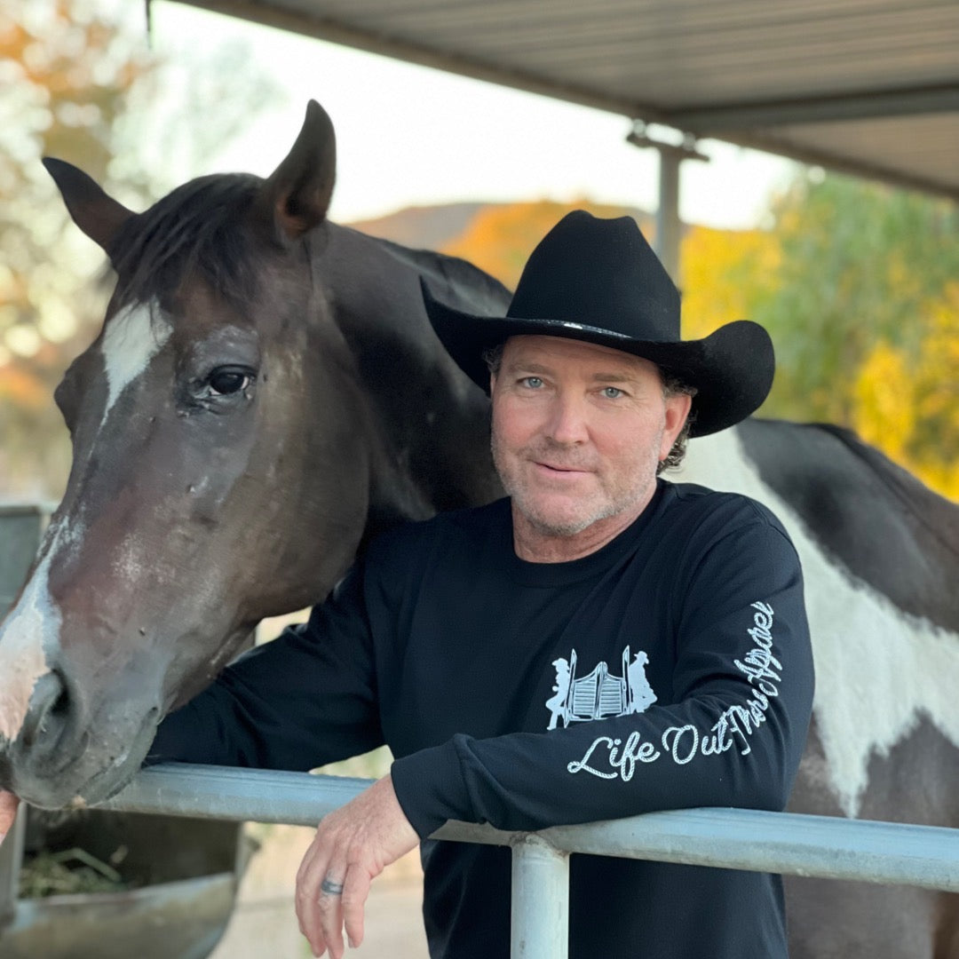 A man in a black "Unisex THE YEEHAW Long Sleeve T Shirt" from Life Out There Apparel and cowboy hat stands next to a black and white horse, embodying classic cowboy style. Set against the backdrop of a ranch, with a metal fence and shelter visible, he rests his arm on the fence, looking directly at the camera.