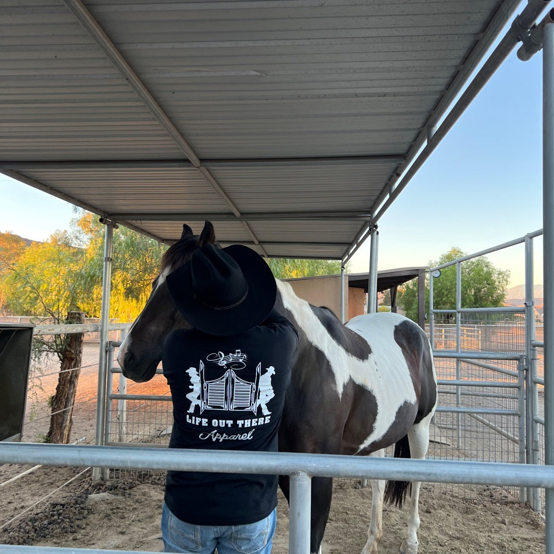 A person exuding cowboy style, wearing a black hat and a Unisex THE YEEHAW Long Sleeve T Shirt - Black / White from Life Out There Apparel, stands with their back to the camera, petting a black and white horse in a covered stable. Trees and a sunset sky hint at the serene ranch life unfolding in the background.