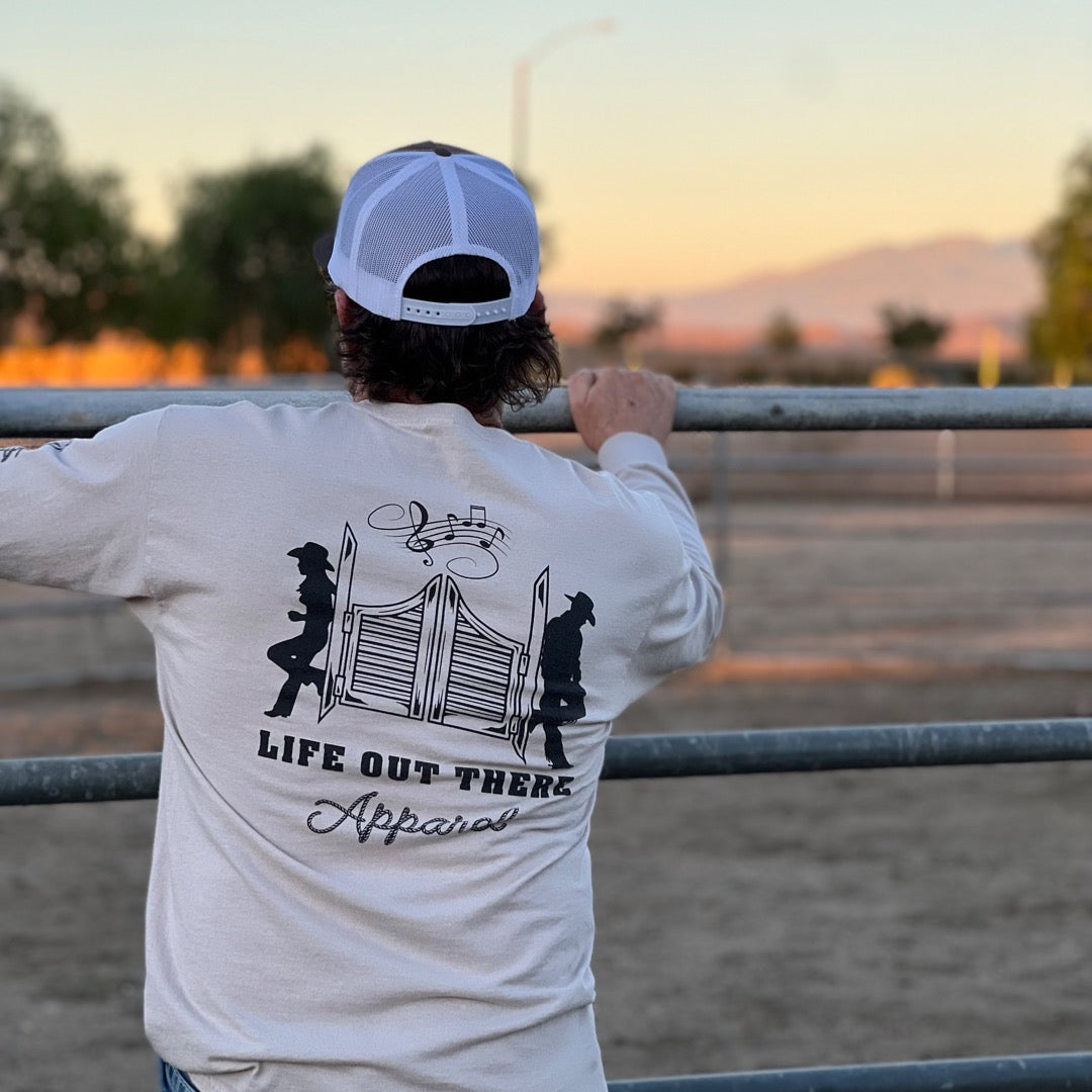 A person dressed in a sandstone and black long-sleeve unisex shirt and cap rests against a metal fence, capturing the essence of ranch life. The shirt displays silhouettes of a cowgirl and cowboy alongside a barn door, accompanied by the text "Life Out There Apparel." In the background, open fields and distant trees further reflect the Yeehaw culture.