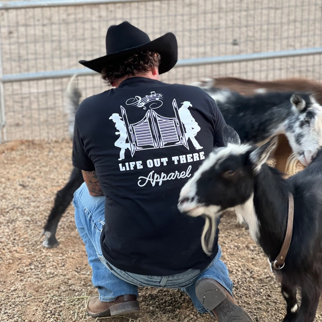 Dressed in classic cowboy attire, including a black hat and Life Out There Apparel's Unisex THE YEEHAW T-Shirt in Black/White, a person kneels beside a group of goats. Set within a fenced outdoor area with gravel underfoot, this scene beautifully captures the essence of ranch life.