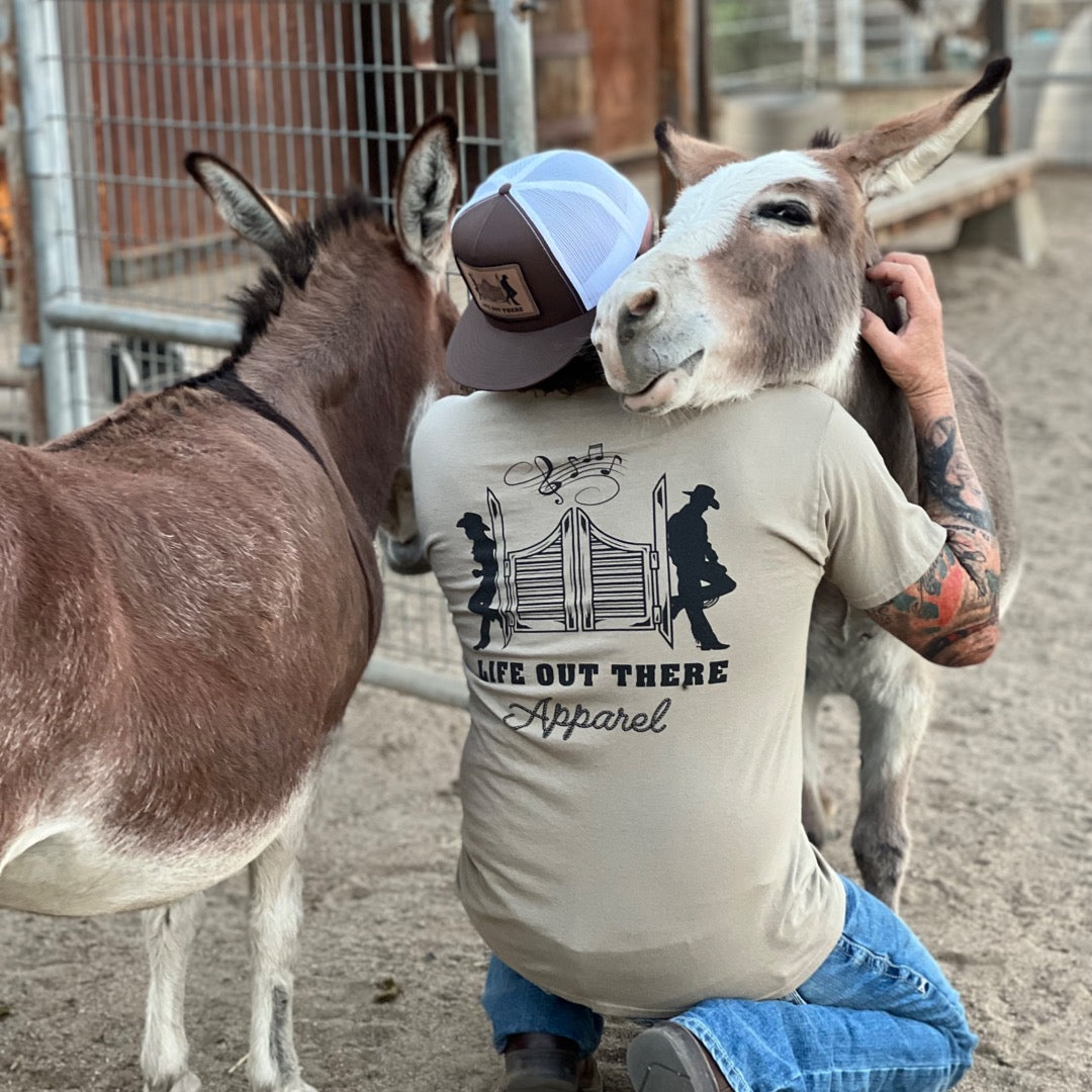A person kneels on the ground, embracing a donkey that rests its head on their shoulder. Clad in a cap and an XXL unisex THE YEEHAW T Shirt by Life Out There Apparel in Sandstone and Black, they evoke the spirit of country life. Nearby, another donkey watches from within a fenced area, as if waiting for its cue in a country music scene.