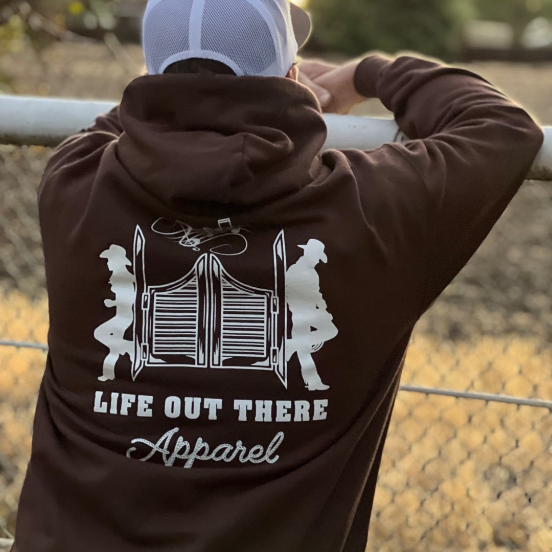 A person in a chocolate and white unisex THE YEEHAW Hoodie from Life Out There Apparel stands facing a chain-link fence, with a blurred natural background that exudes an earthy vibe, capturing the spirit of cowboy culture and ranch life.