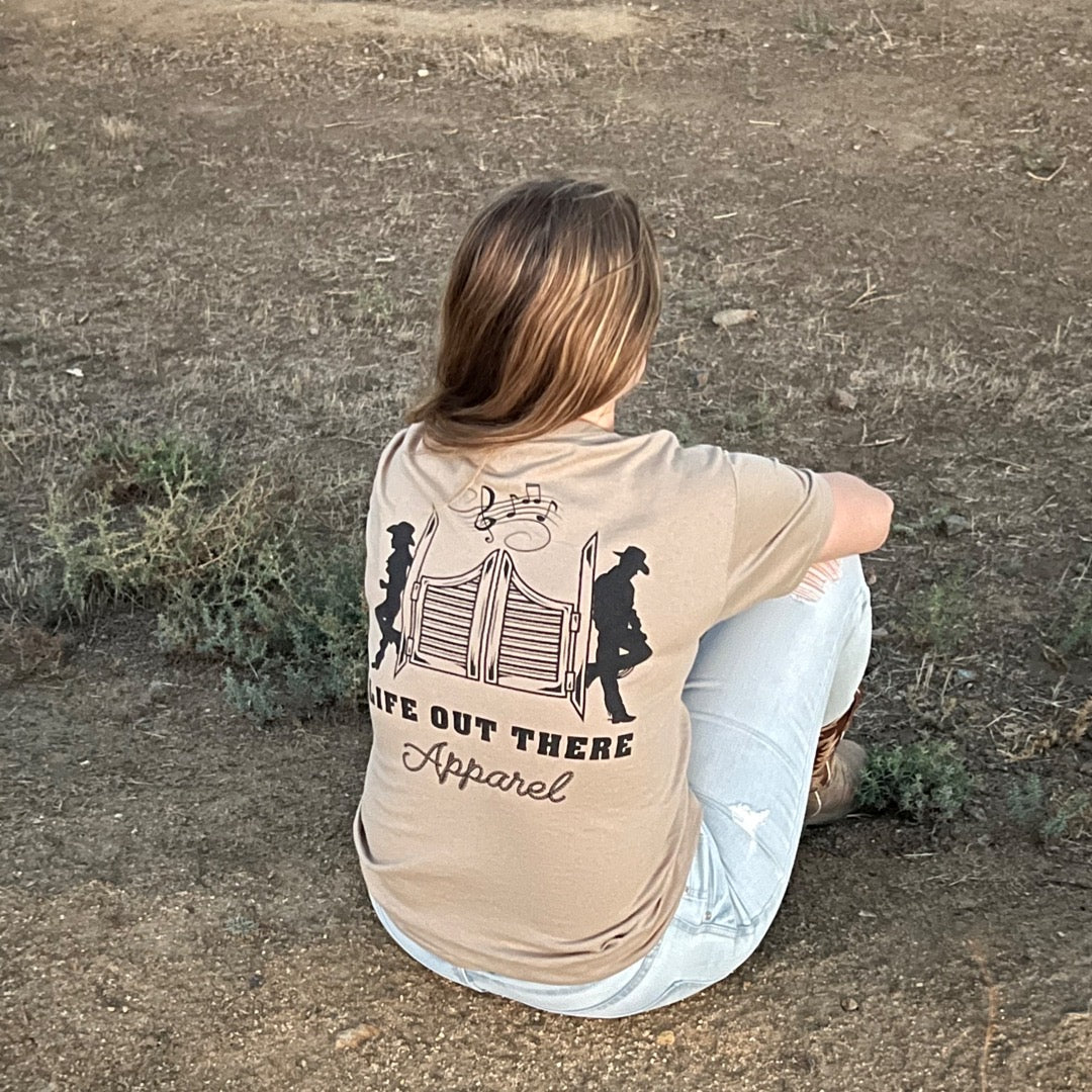 A person with long hair sits on the ground facing away, wearing the Unisex THE YEEHAW T Shirt in sandstone and black from Life Out There Apparel. The dry, scarcely vegetated ground evokes a cowboy style vibe reminiscent of ranch life.
