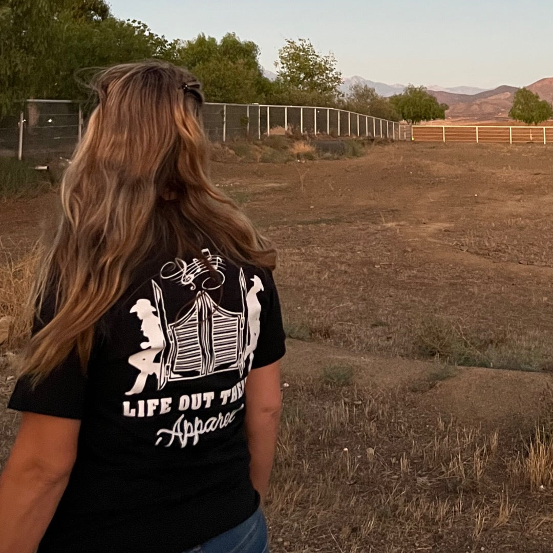 A person sporting the Unisex THE YEEHAW T Shirt in black and white by Life Out There Apparel stands in an open field, encapsulating the essence of ranch life. The T-shirt showcases a graphic design on the back. The setting features trees, a fence, and distant hills beneath a clear sky.