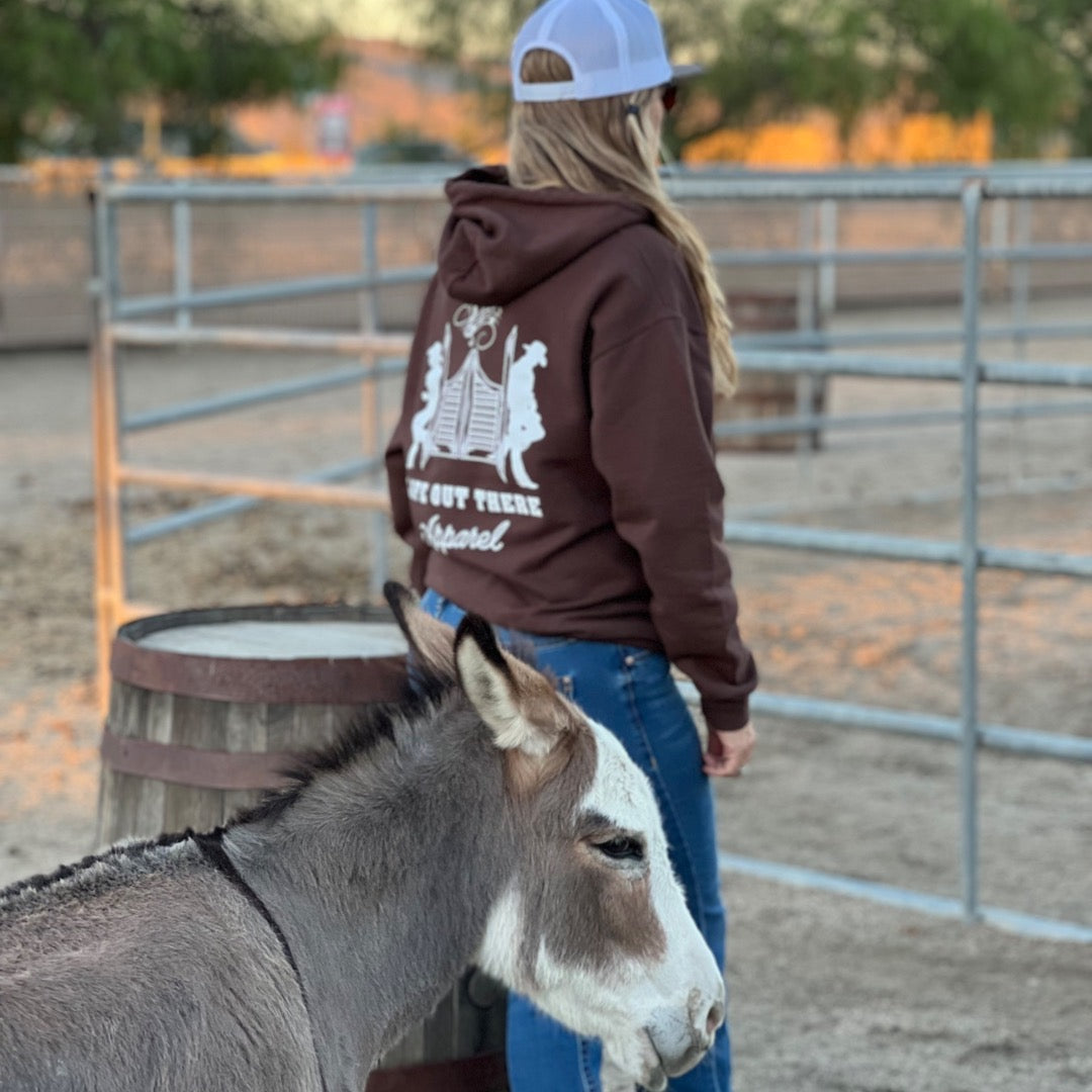 Sporting the Unisex THE YEEHAW Hoodie in chocolate and white by Life Out There Apparel, a person channels a ranch-life vibe. They stand beside a donkey within a fenced area, with distinct logo and text detailing on the hoodie. In the background, two wooden barrels and trees enhance the rustic charm of the scene.