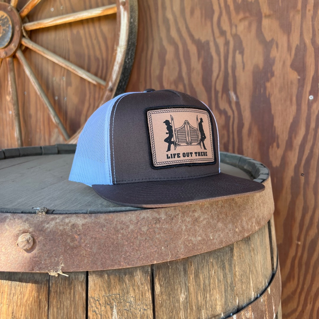 A brown and white "Life Out There - THE YEEHAW" SnapBack Trucker Hat by Life Out There Apparel, featuring a brown patch with silhouettes of people lifting a log and the words "Life Out There," rests on a wooden barrel beside a rustic wooden wheel.