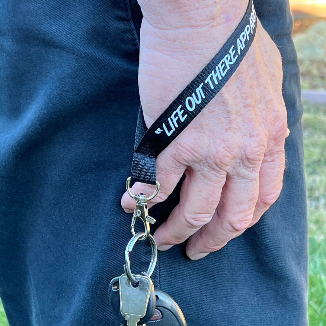 A person holding keys attached to a wrist lanyard keychain from Life Out There Apparel, which features the brand name, is standing outdoors on grass.