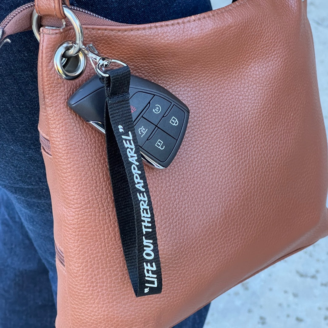 A chic brown leather handbag elegantly drapes from a Life Out There - Wrist Lanyard Keychain by Life Out There Apparel, featuring a car key fob on a sleek black strap. This stylish accessory is casually held against someone in blue jeans, seamlessly merging fashion with function.
