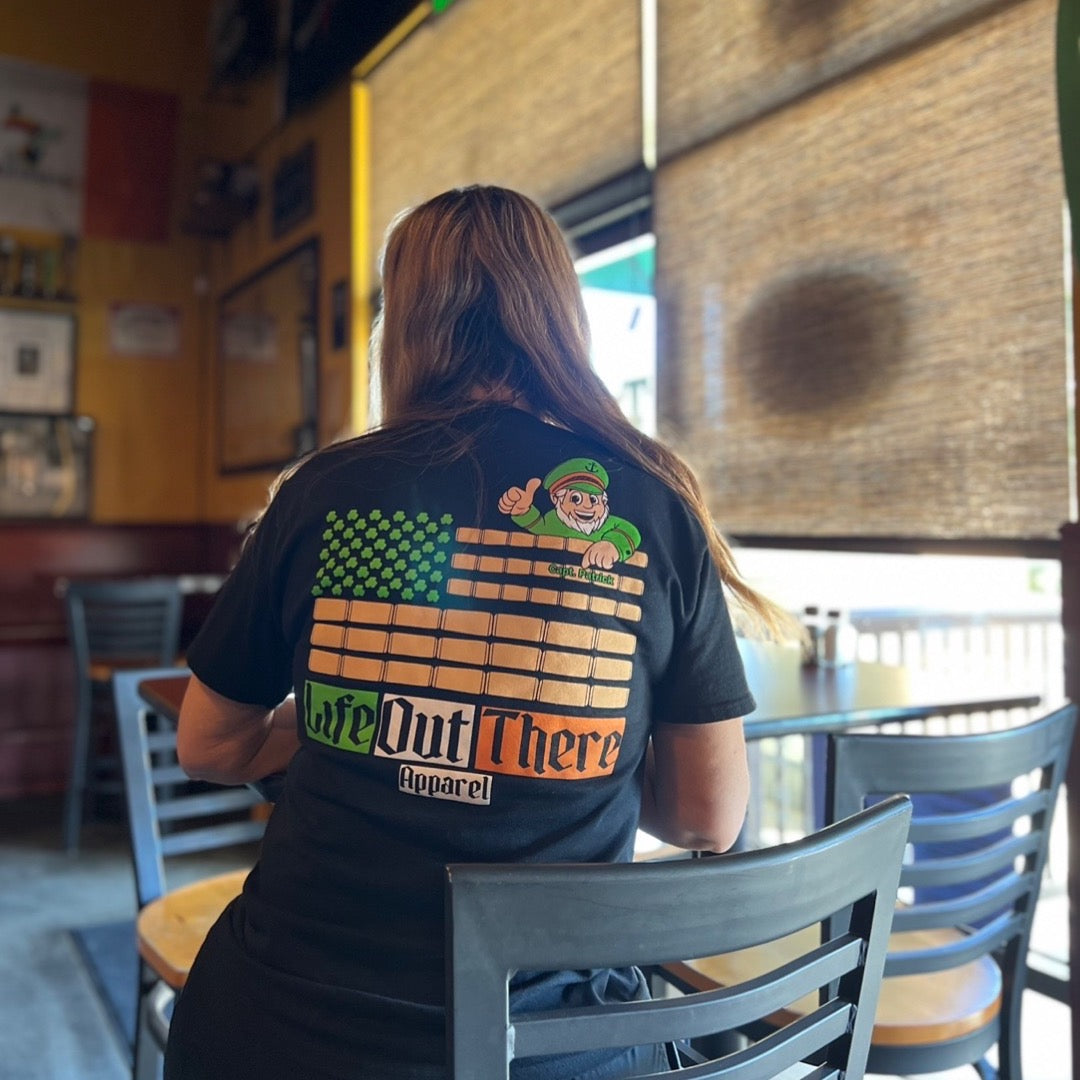 Life Out There Apparel “Capt. Patrick” Unisex tee shirt in black. This is a collaboration of an American flag with an Irish twist and a leprechaun boat Captain looking on. This photo has a women sitting in a pub.