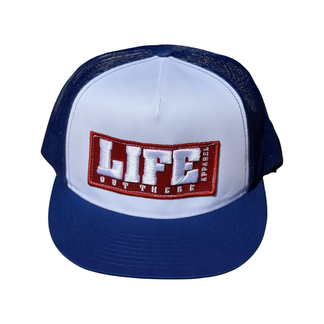 The Life Out There - Puff Embroidered SnapBack Trucker Hat from Life Out There Apparel is a royal blue and white trucker hat with a mesh back. It features the brand's bold logo on the front, showcasing "LIFE APPAREL" in prominent white letters set against a red background, accompanied by "OUT THERE" in smaller red letters on a white backdrop below.