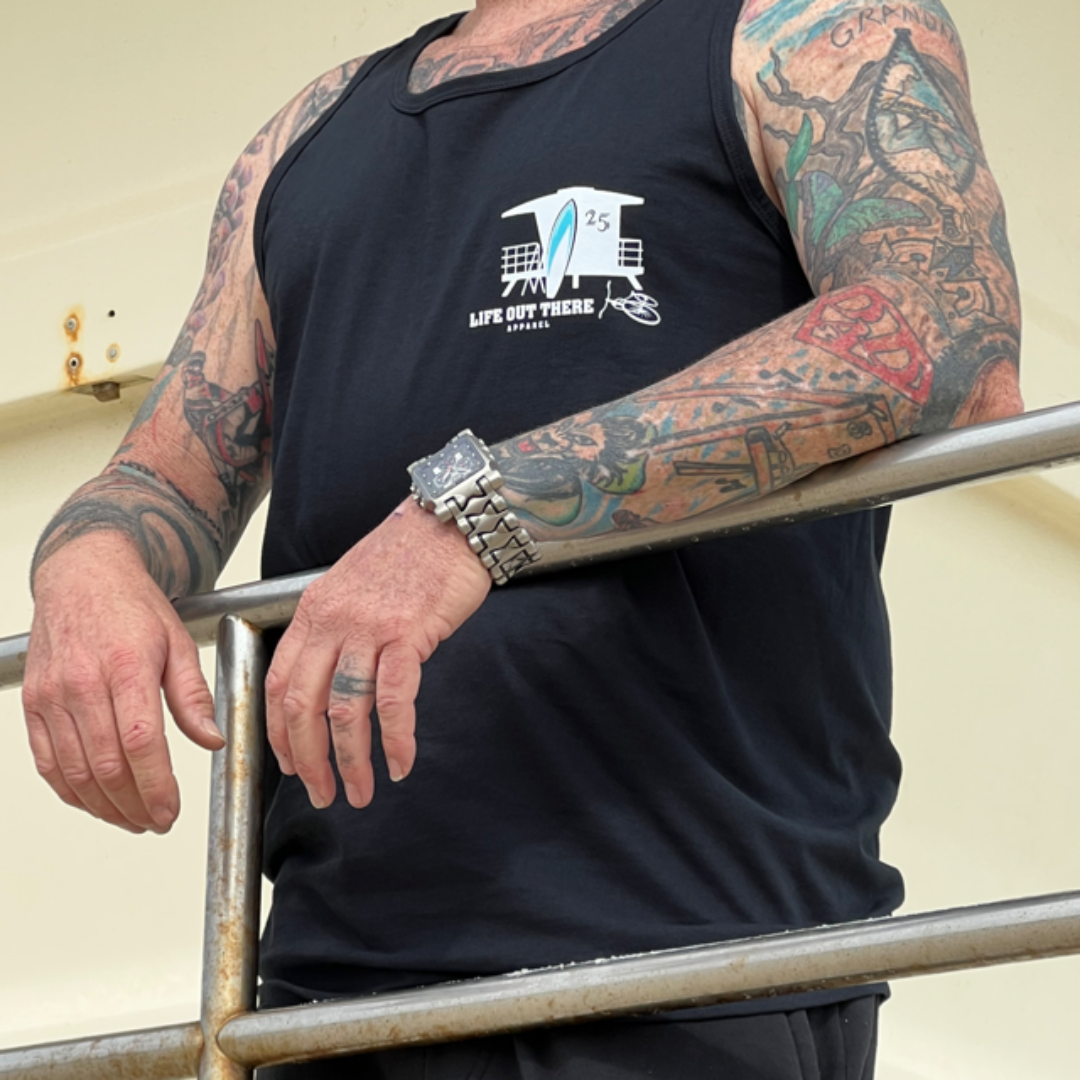 A person with tattooed arms leans on a railing, wearing the Unisex The JJ Tank Top in black by Life Out There Apparel, which features an ocean-themed design. They also wear a watch, showcasing their upper body and tattoos as if they're embracing the cool vibe of surfing near a lifeguard tower.