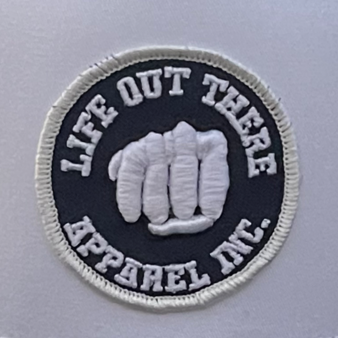 The centerpiece of the circular patch is a striking white fist design, accompanied by "Life Out There Apparel" text, set against a stylish black backdrop. Enclosed by a crisp white border, this emblem makes an eye-catching addition to any navy SnapBack or trucker hat collection.
