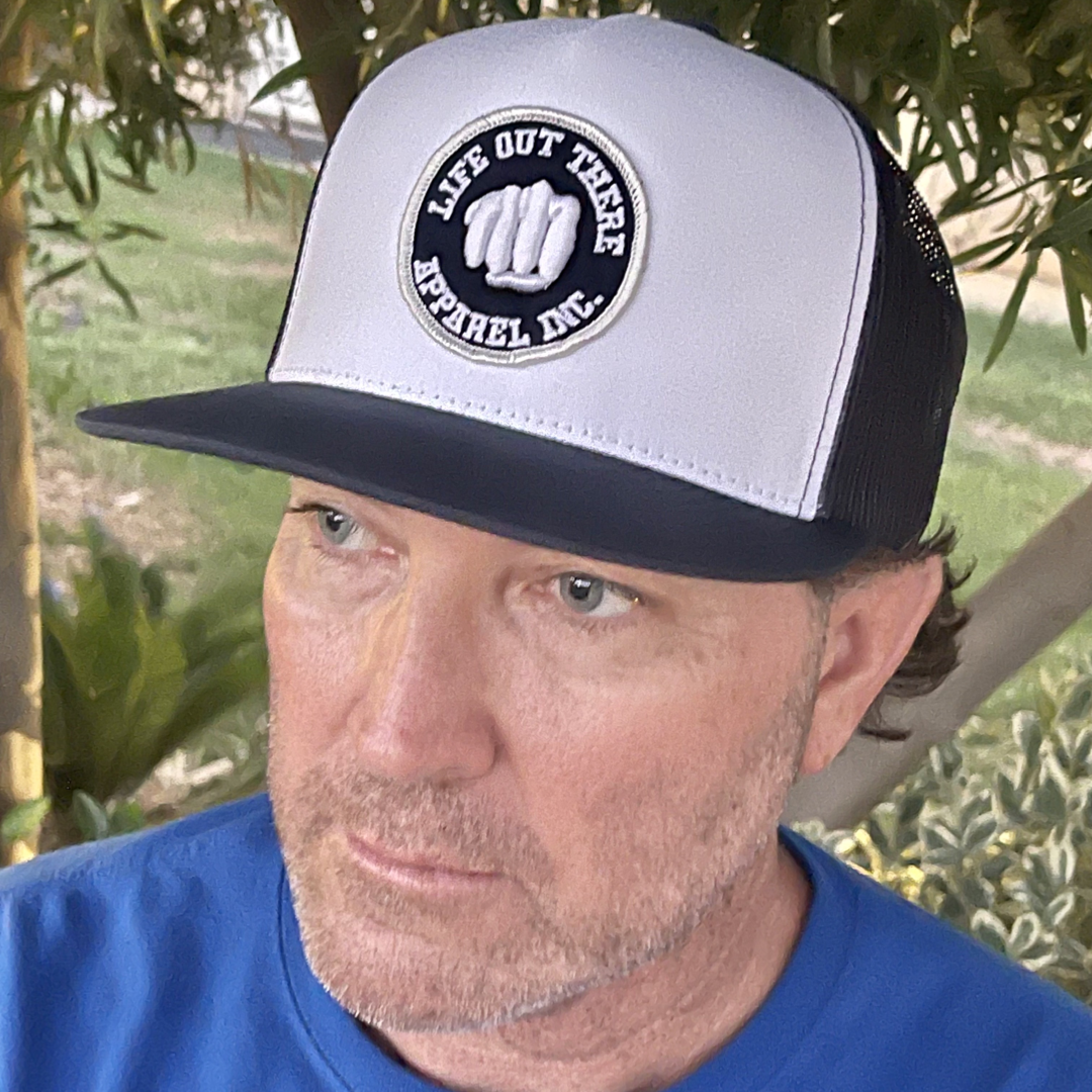 A man with a beard wears a navy blue shirt and The Pound It - 3D Puff Embroidered Snapback Trucker hat in navy/white from Life Out There Apparel. The backdrop is a lush garden filled with green plants and a towering tree.