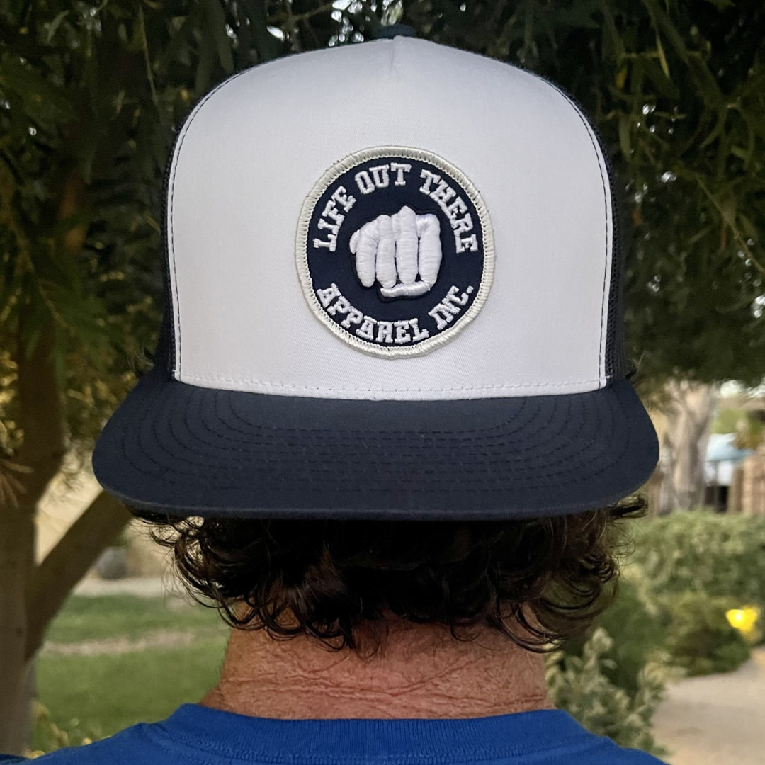 A person wearing "The Pound It - 3D Puff Embroidered Snapback Trucker - Navy/White" by Life Out There Apparel, featuring a black and white front logo with a stylized fist, against a background of blurred greenery that accentuates the hat's distinctive design.