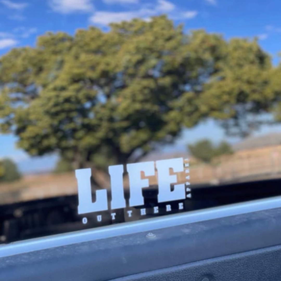 The image displays the "Life Out There - Die Cut Sticker - 8 inch" from Life Out There Apparel, adhered to a car window. The sticker reads "LIFE OUT THERE" in prominent text with "ISNT ANYWHERE" beneath it in smaller lettering. Reflected in the window are a large tree and a blue sky with clouds, enhancing the serene outdoor ambiance.