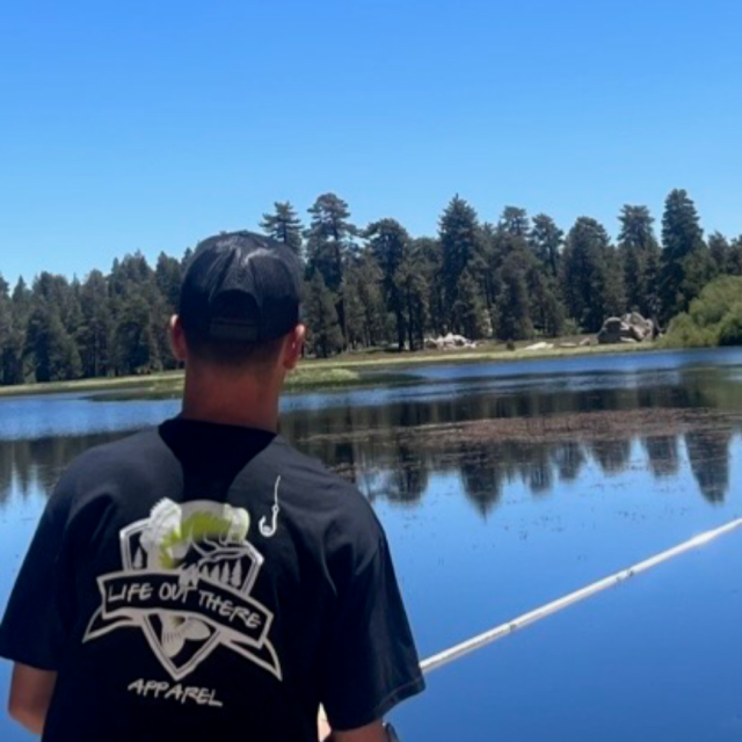 A person dressed in a **Life Out There Apparel's Unisex The Hooker T Shirt** with "THE HOOKER" logo on the back is fishing at a tranquil lake under a clear blue sky. A shimmering gold fish hook adds sparkle to the peaceful scene, surrounded by dense trees.