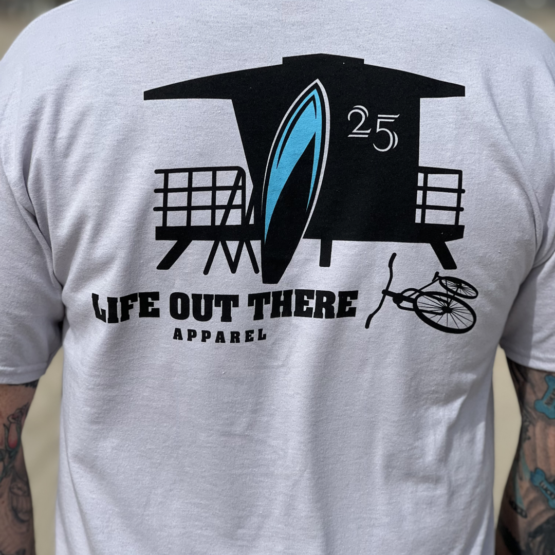 A person wearing a silver JJ Tee from Life Out There Apparel, which showcases a graphic of a lifeguard tower, surfboard, and bicycle at Huntington Beach with the number 25. The person's arms display visible tattoos.