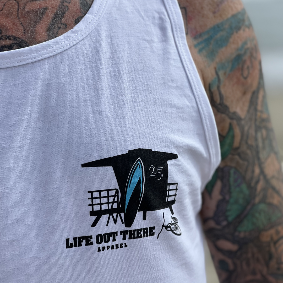 A person wearing the Unisex The JJ Tank Top - White by Life Out There Apparel, featuring a lifeguard tower design with a surfboard, perfectly capturing Huntington Beach vibes. Their tattooed arm vividly reflects the essence of surfing spirit.