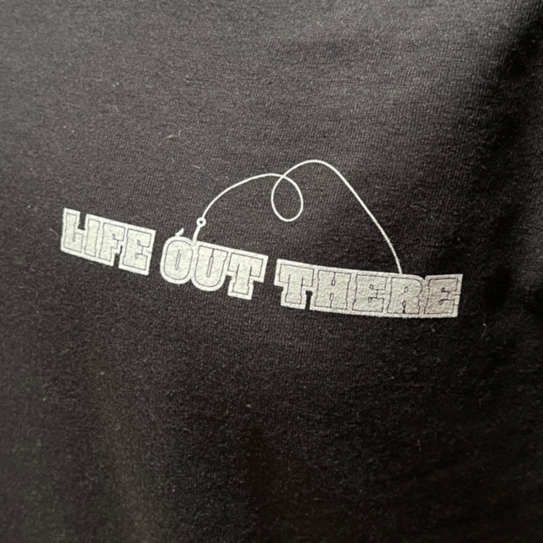 The image features the Unisex The Hooker T Shirt in black and white from Life Out There Apparel. It showcases the text "LIFE OUT THERE" in a blocky font, complemented by a sleek line above that mimics a fishing line or gold fish hook, contributing to its minimalist design.