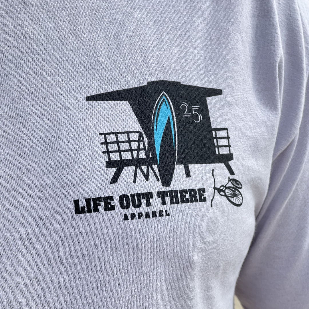 A close-up of the Unisex The JJ Tee in Silver showcases a lifeguard tower and surfboard graphic, embodying beach life with Life Out There Apparel. A bicycle silhouette enhances the coastal vibe, reminiscent of relaxing days in Huntington Beach.