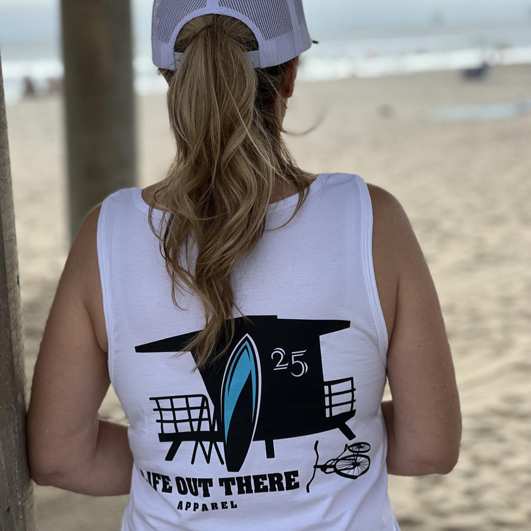 Wearing a white "Unisex The JJ Tank Top" by Life Out There Apparel, a person with long hair styled in a ponytail stands near Huntington Beach. The tank features a lifeguard tower, surfboard, and the number 25 on the back, as they enjoy the view of sand and ocean creating perfect surfing waves.