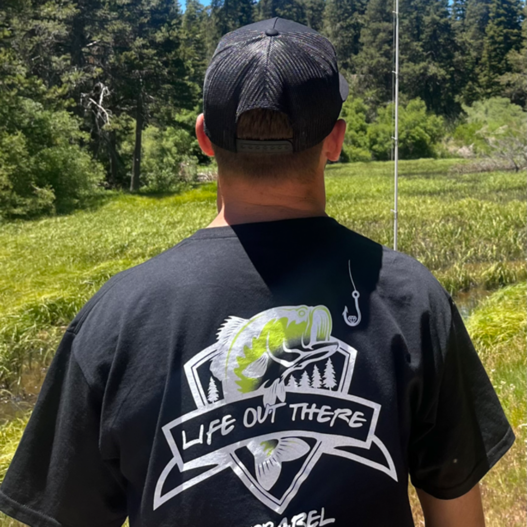 A person wearing a black Unisex The Hooker T Shirt by Life Out There Apparel, showcasing a large fish and trees design, is standing in a lush green field facing away. To the right, there's a fishing rod with a glimmering gold fish hook set against the backdrop of an enchanting forest.