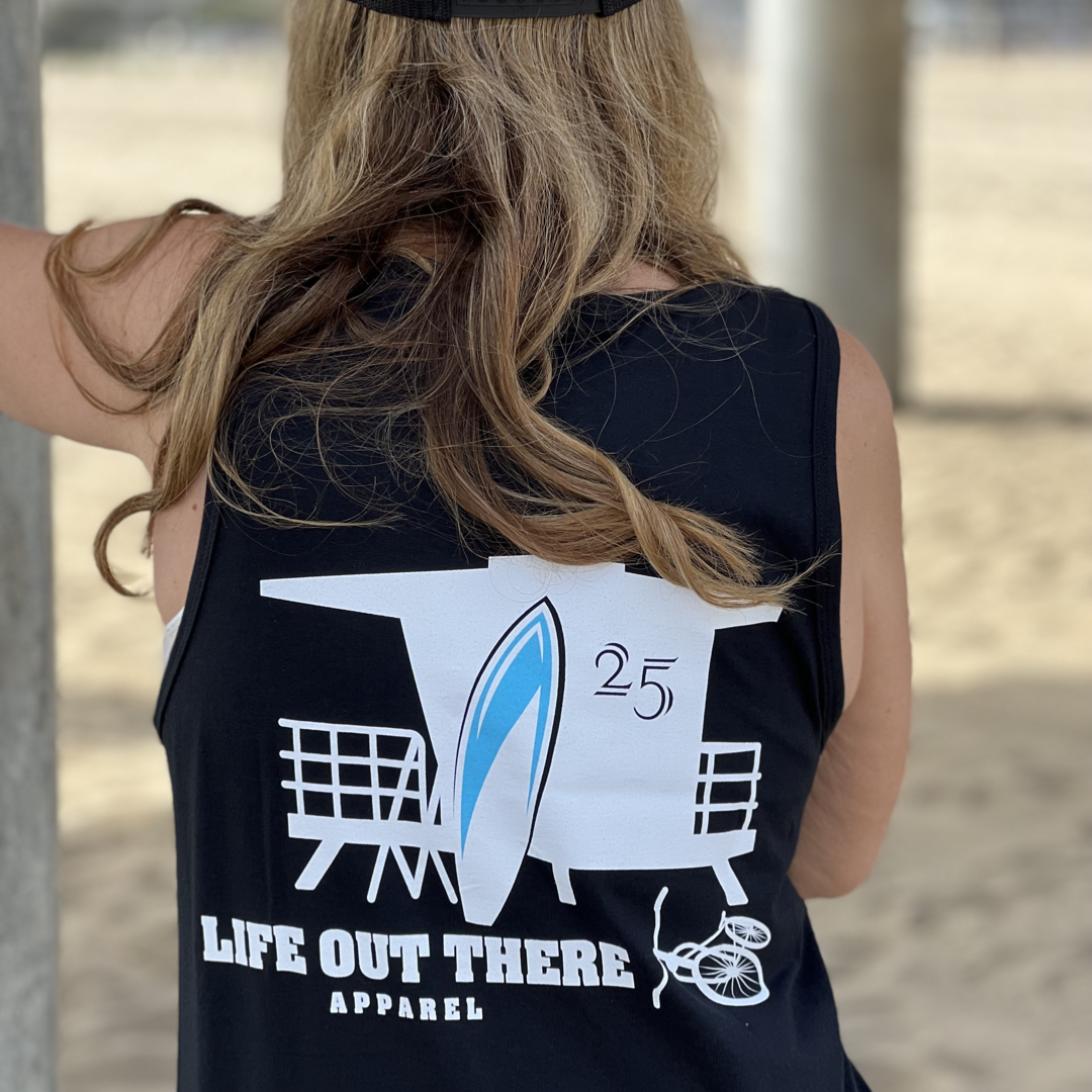 A person is wearing the Unisex The JJ Tank Top in black, featuring a lifeguard tower, surfboard, and the number 25. Below this design, the brand name "Life Out There Apparel" is inscribed. The sandy background suggests the lively surfing culture of Huntington Beach.