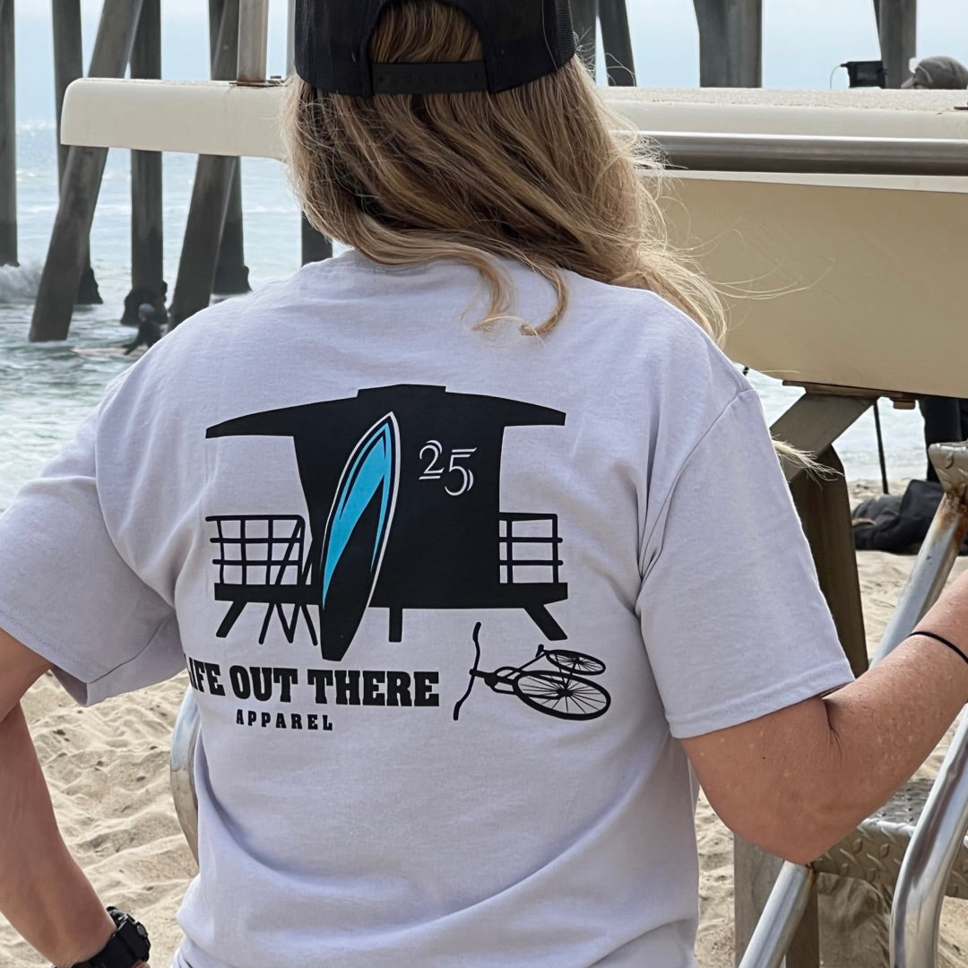 A person wearing the Unisex The JJ Tee in Silver by Life Out There Apparel, featuring a graphic of a lifeguard tower and surfboard, stands on Huntington Beach near a pier with their surfboard resting beside them.
